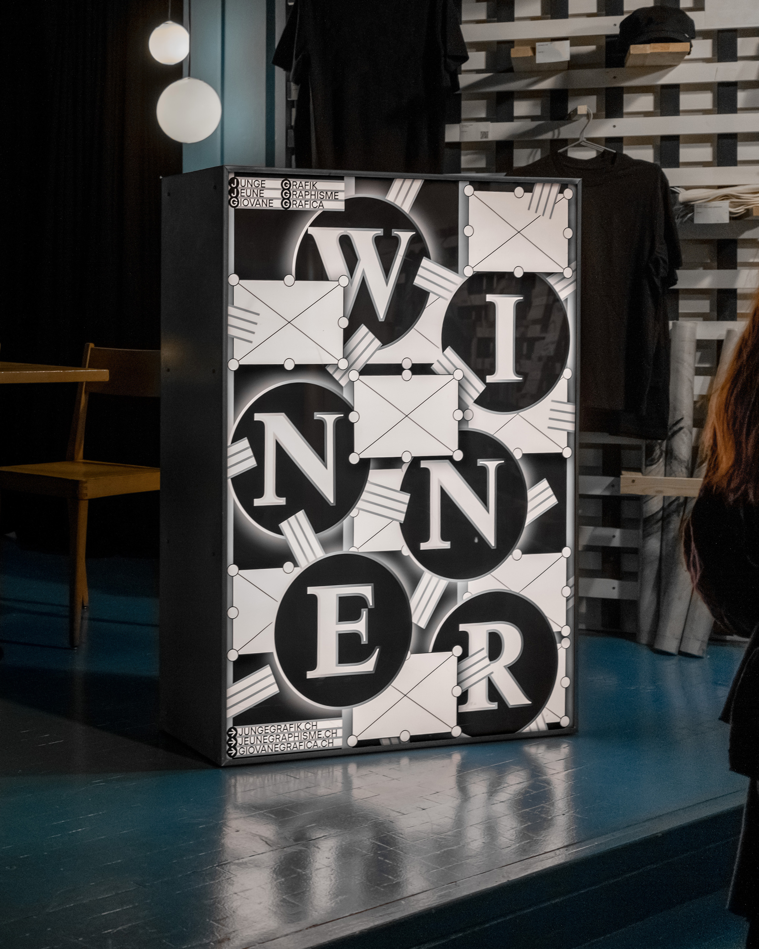 Illuminated display box with letters spelling WINNER in large black circles, set in an indoor space with chairs and hanging lights in the background.