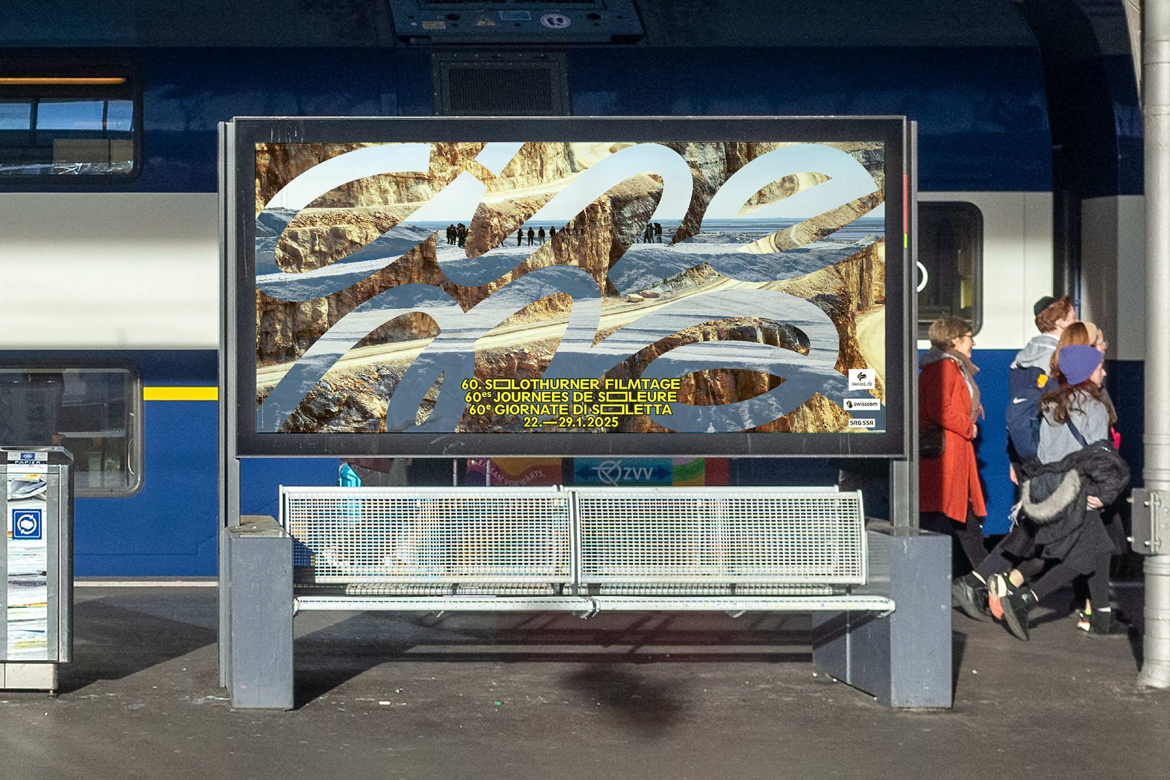 Outdoor train station platform with a metal bench in front of a large illuminated advertisement showing a rocky landscape and event text for the 60th Solothurn Film Festival dated January 22-29, 2025.