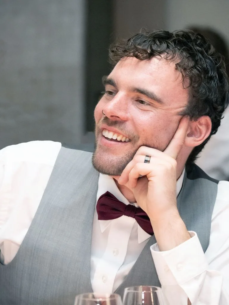 Young man in a white shirt, gray vest, and burgundy bow tie smiling and resting his head on his hand.