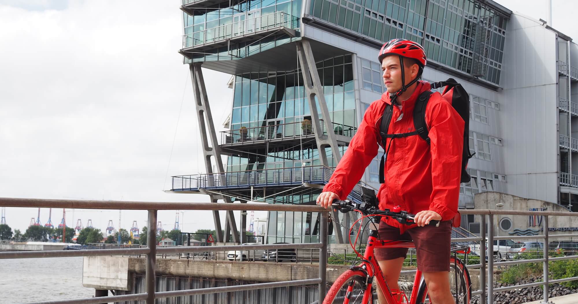 A cyclist wearing a red helmet, red jacket, and backpack stands with a red bicycle on a riverside walkway in front of a modern glass building. The scene appears urban and contemporary, with industrial cranes visible in the distant background across the river.