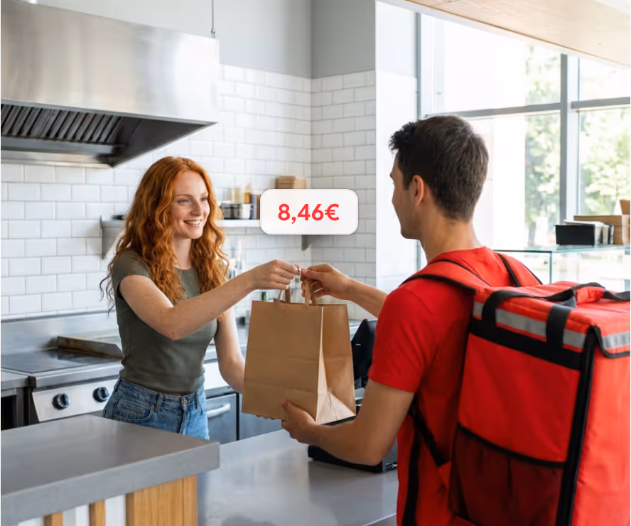 A woman behind a counter hands a paper bag to a delivery person wearing a red shirt and backpack with a price tag of 8,46€ visible.