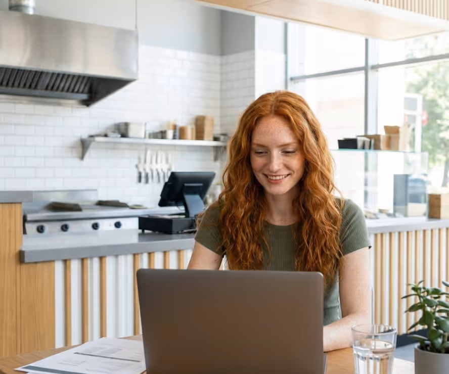Smiling woman with long red hair working on a laptop at a table in a bright modern restaurant kitchen.