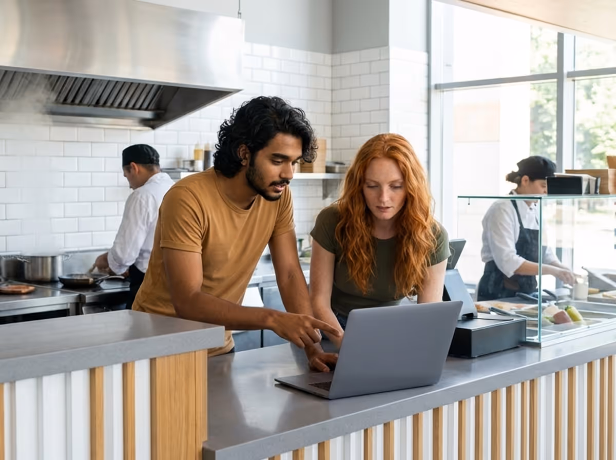 Man and woman looking at a laptop together across a counter in a modern kitchen with chefs cooking in the background.