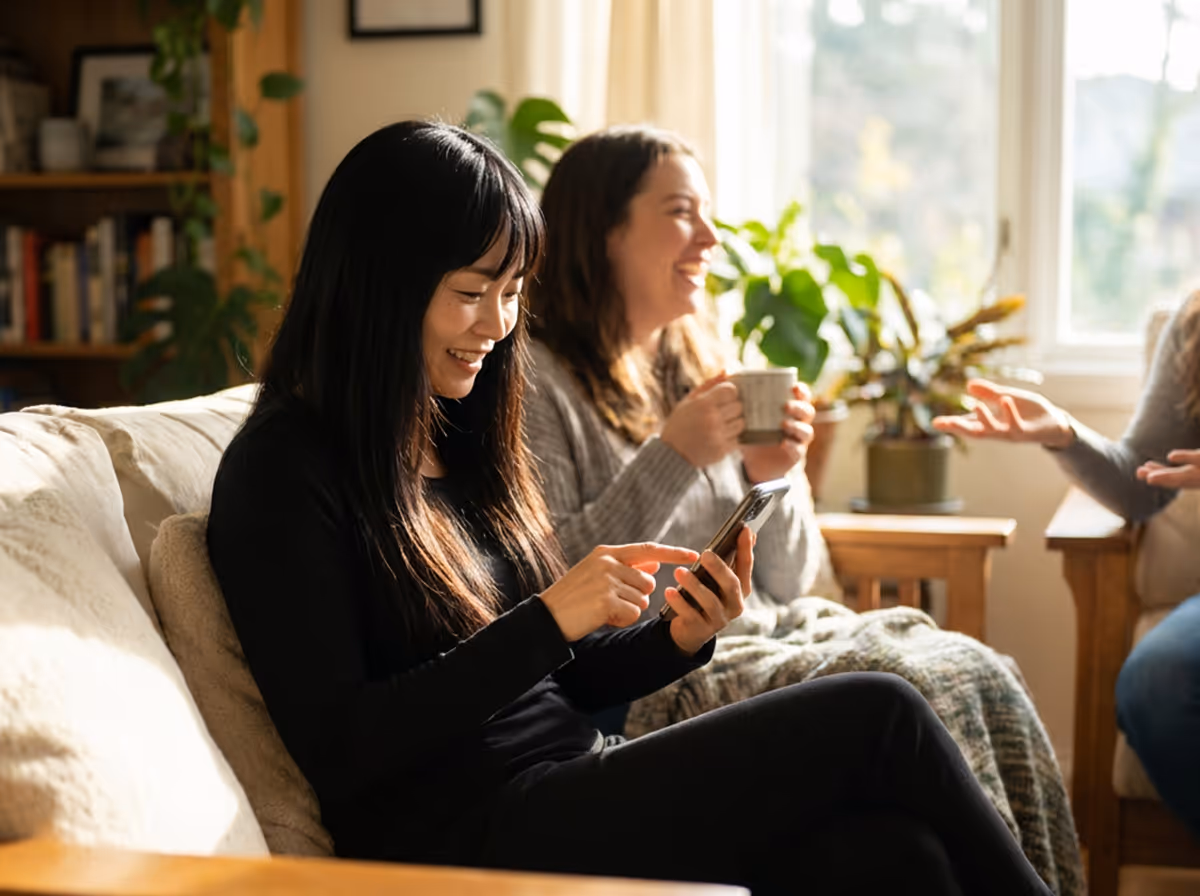 Two women sitting on a couch, one smiling and using a smartphone, the other holding a mug and laughing, in a sunlit room with plants.