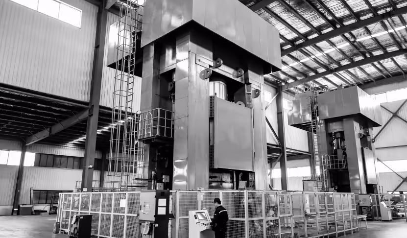Two large industrial forging machines inside a metal factory, with a worker operating a control panel in the foreground.