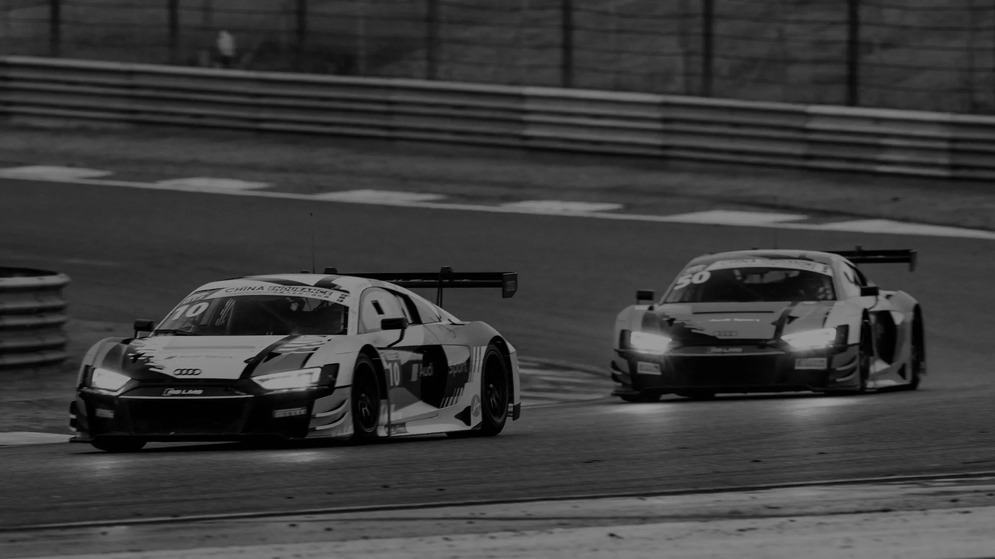 Two Audi race cars with headlights on navigating a curve on a race track.