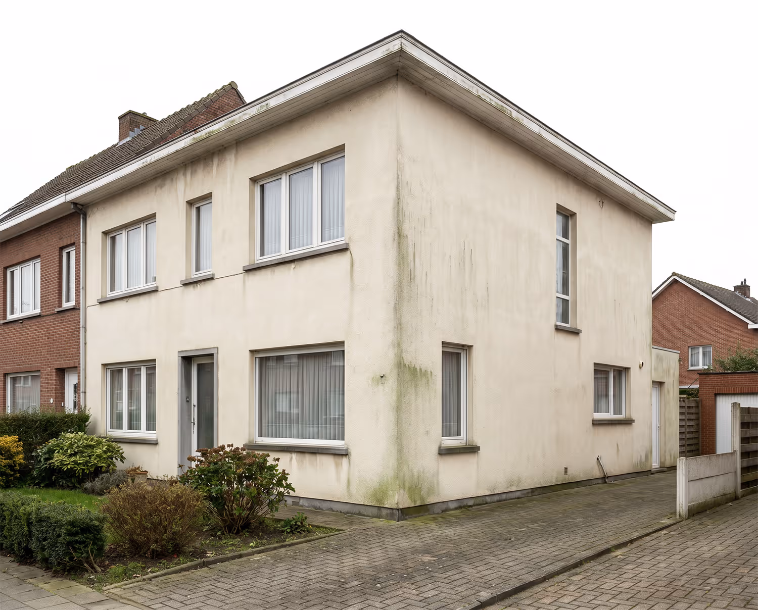 Woonhuis met een beige gevel met zichtbare groene aanslag en waterstrepen naast een straat met bestrating.