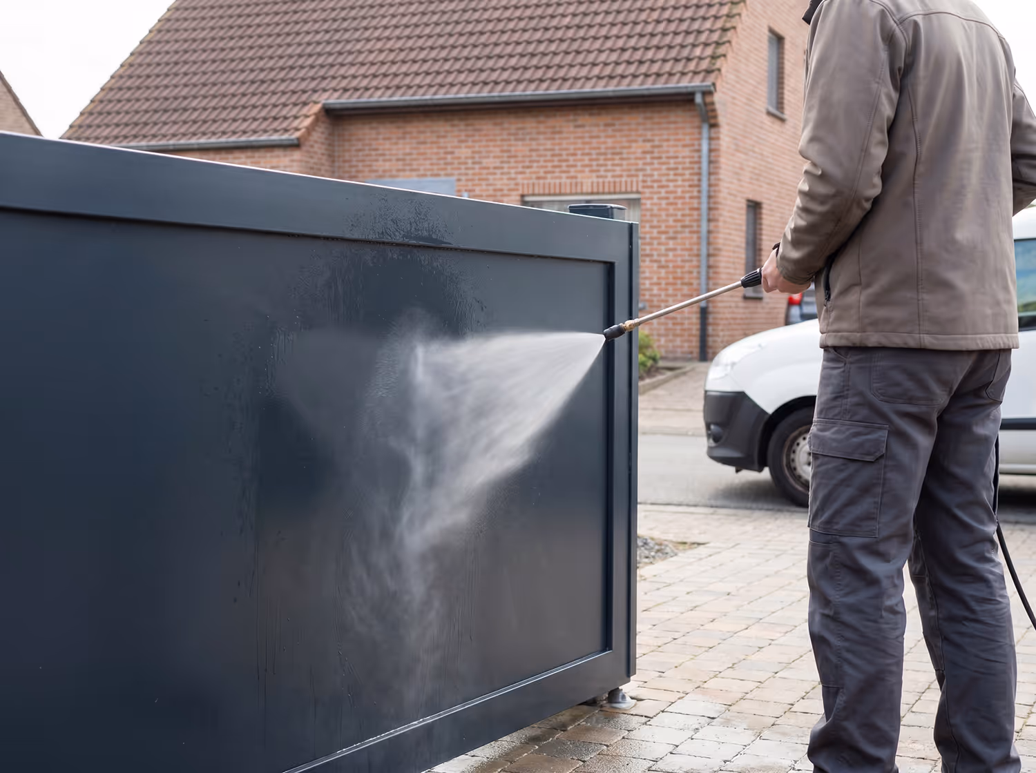 Man die met een softwash reiniger een donkere metalen poort schoonspuit in een woonwijk.