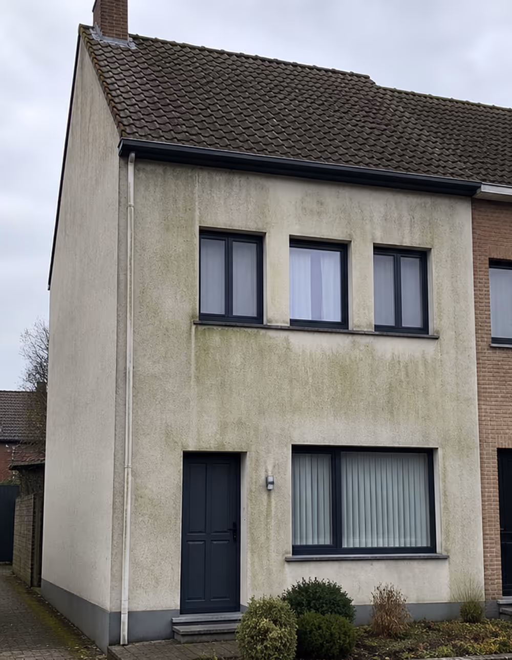 Twee verdiepingen tellend huis met hoekdak, groenige en bevuilde gevel en zwarte ramen en deur.