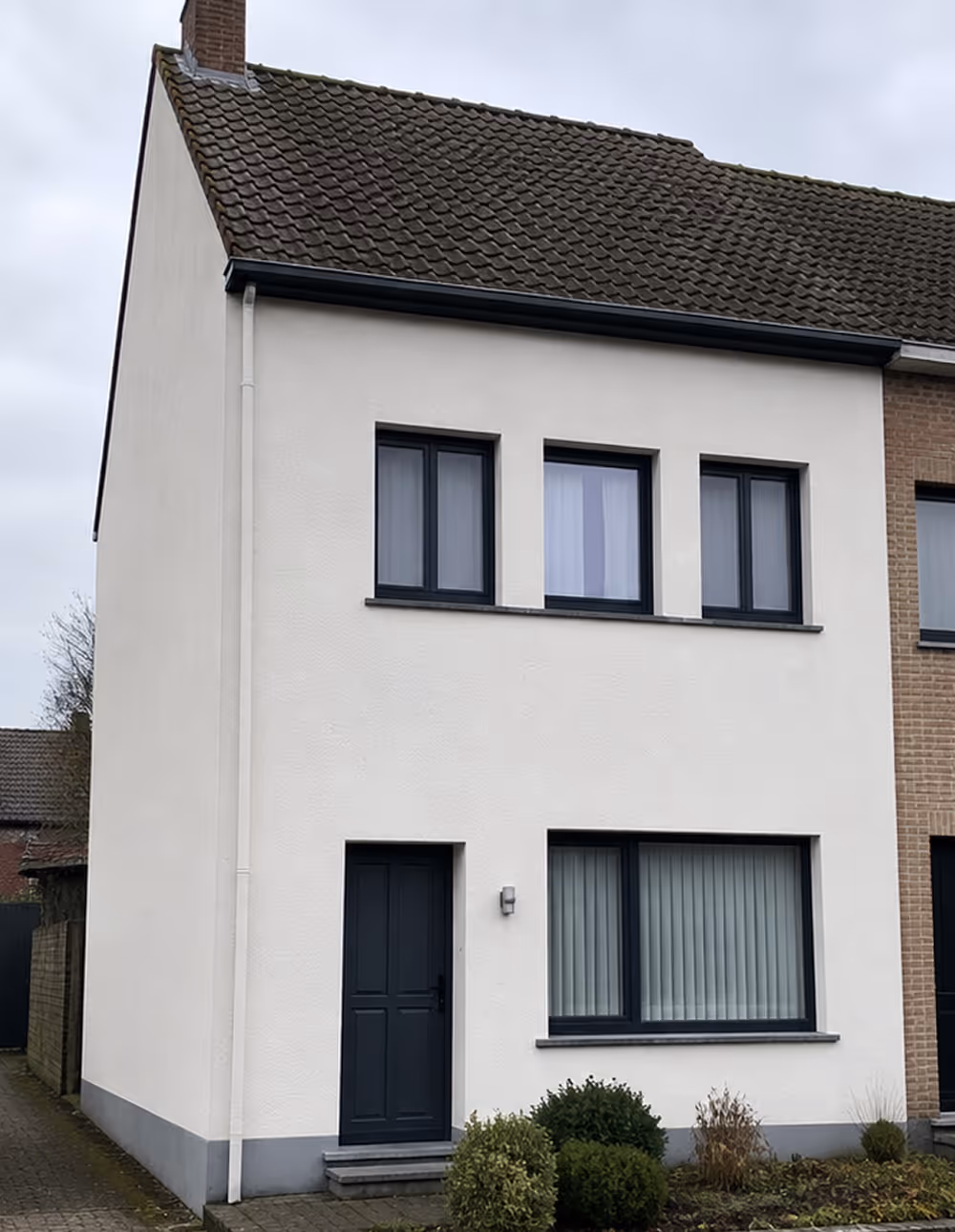 Eengezinswoning met witte geverfde voorgevel, donkergrijze deur en vensters, en roodbruine dakpannen.