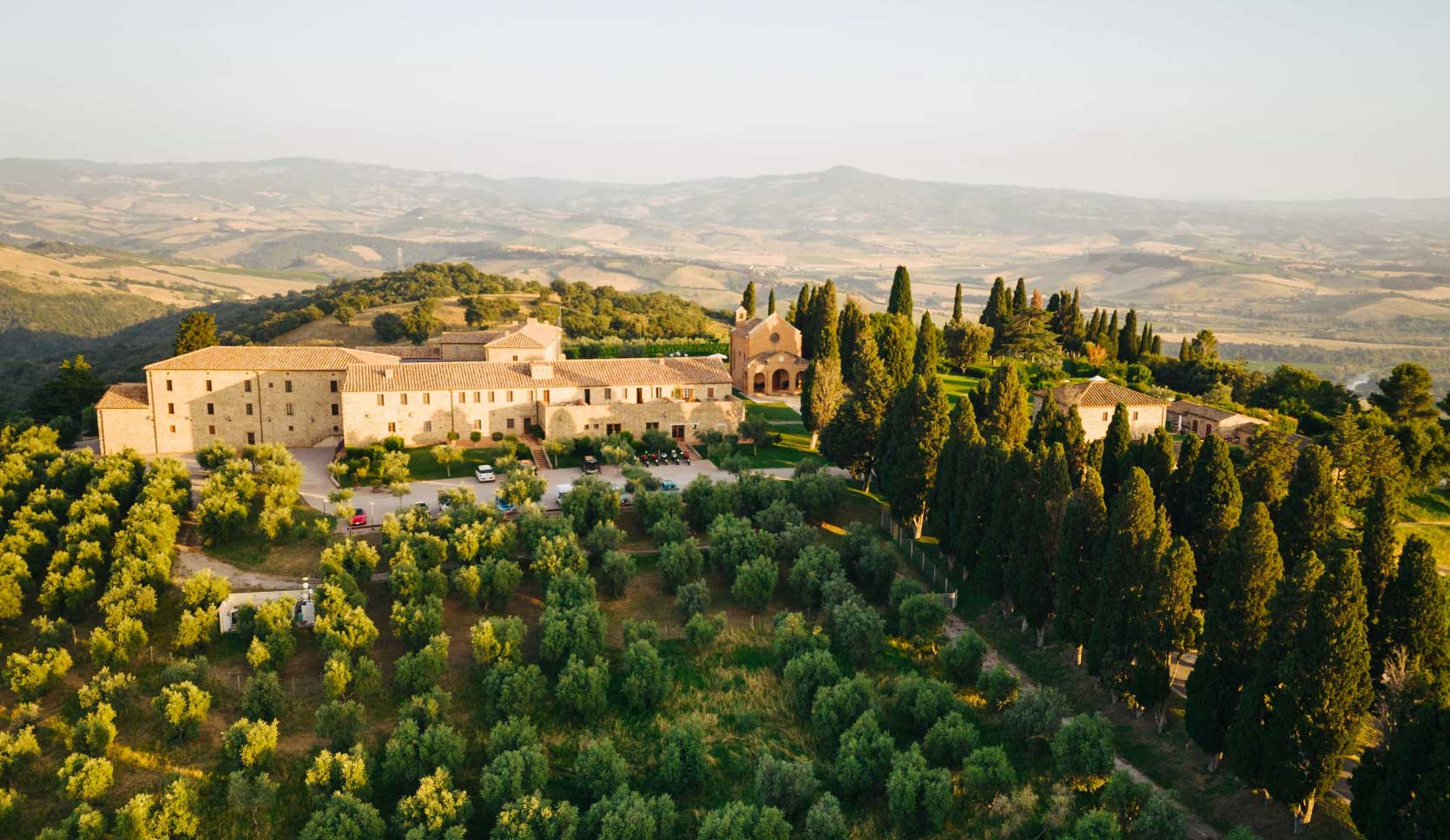 Aerial view of ColleMassari Estate surrounded by olive groves and tall cypress trees in a hilly countryside during golden hour.