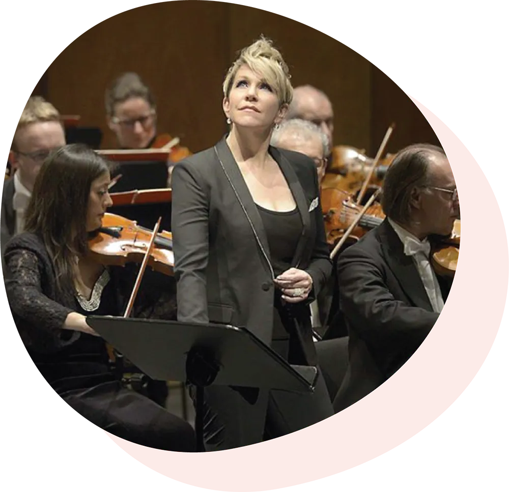 Joyce DiDonato in a black suit standing before an orchestra with violinists behind her.