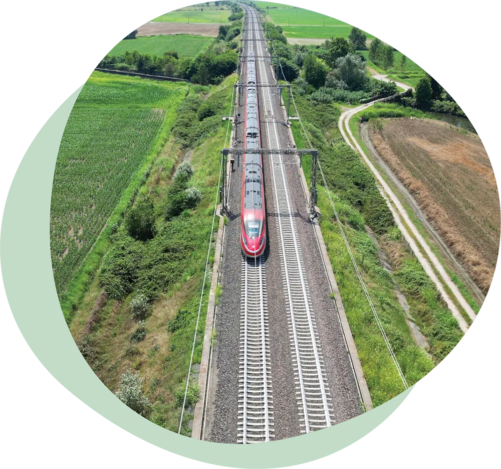 A red high-speed train traveling on electrified railway tracks through green fields and farmland in Tuscany region, Italy.