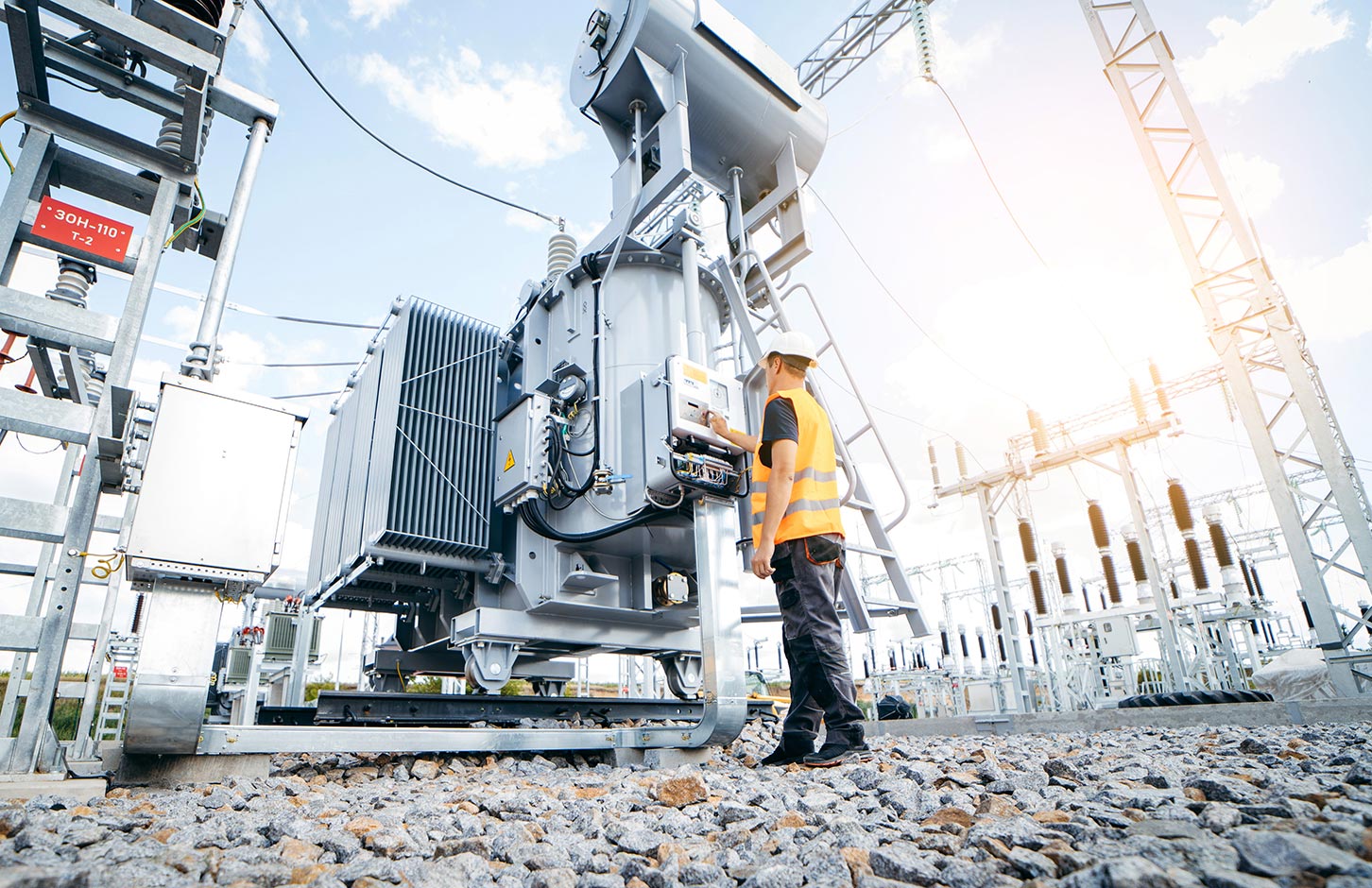 Electrician in a safety vest and helmet inspecting equipment at an electrical substation on a rocky ground under a partly cloudy sky.