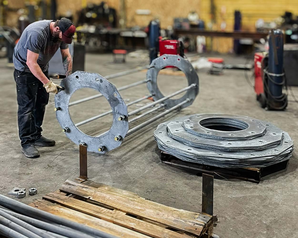 Man in gloves and cap working in a workshop with large metal anchor bolt assembly and stacked metal plates on the floor.