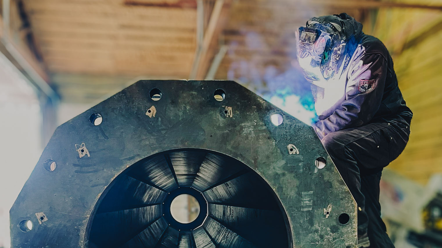 Welder in protective gear working on a large industrial metal piece in a workshop.