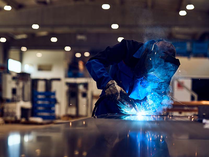 Worker wearing protective gear welding metal in a factory with bright blue sparks and smoke.