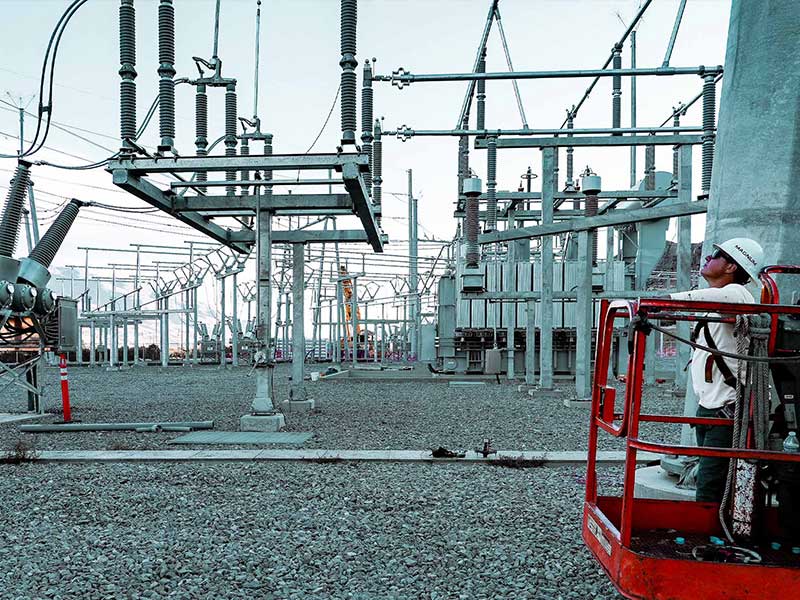 Worker wearing a hard hat inspecting equipment inside an electrical substation with steel structures and insulators.