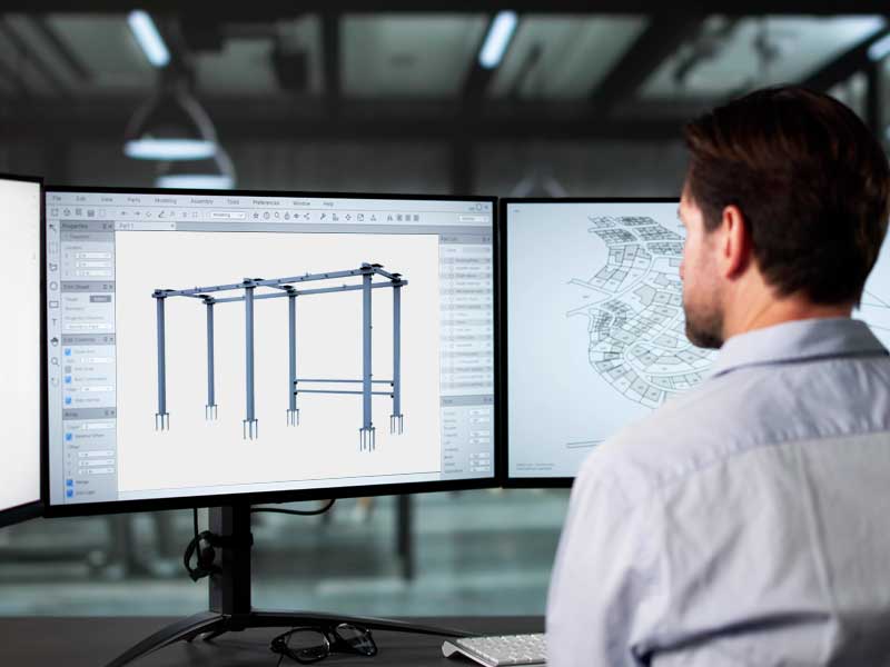 Man in a light shirt sitting at a desk, looking at two large computer monitors displaying a 3D structural model and architectural floor plans.