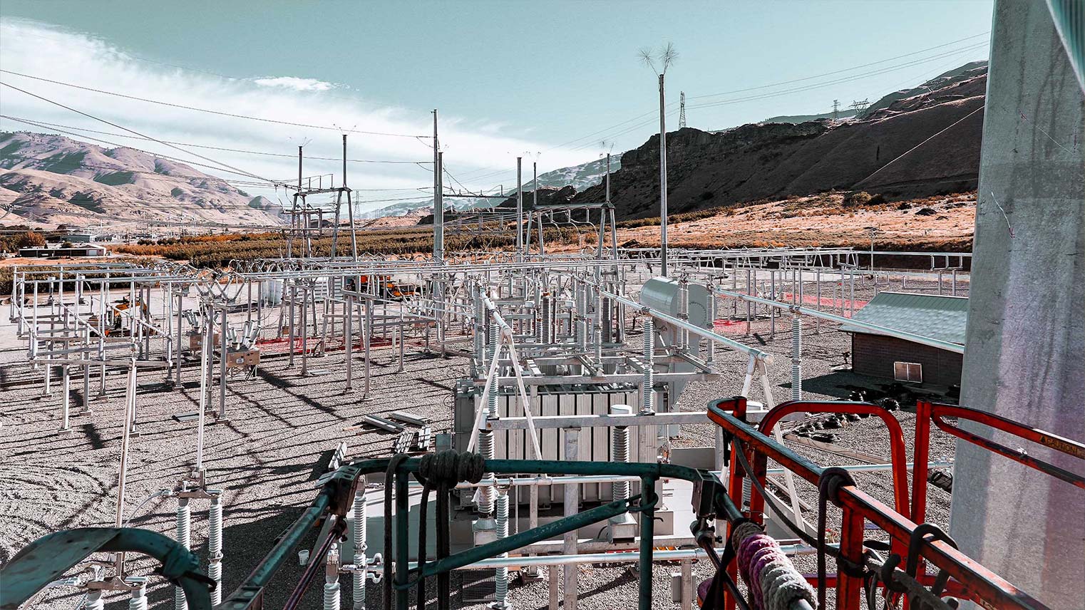 Electrical substation with metal frameworks, transformers, and power lines set in a mountainous landscape under a blue sky.
