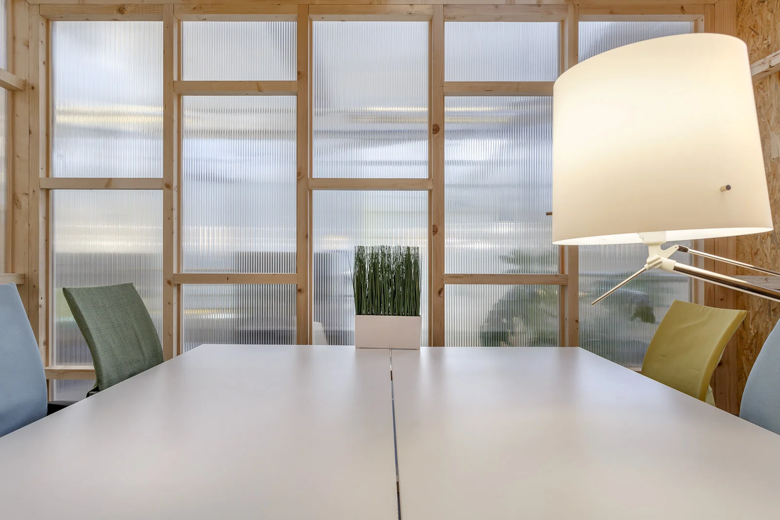 Salle de réunion moderne avec table blanche, chaises colorées, lampe sur pied et paroi vitrée à cadre en bois.