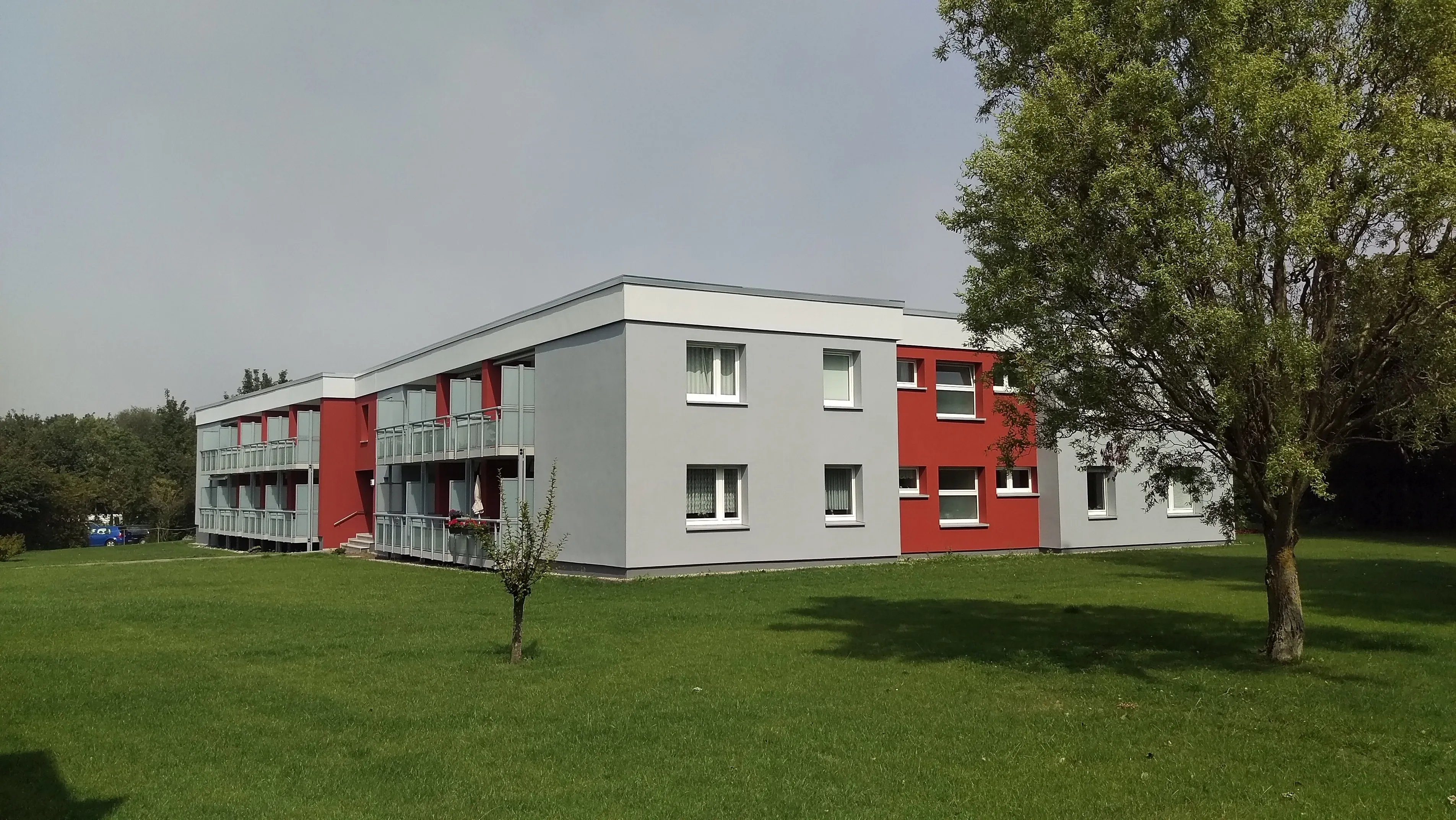 Modernes zweistöckiges Gebäude mit grauen und roten Wänden, angrenzend an eine Grünfläche mit Bäumen.