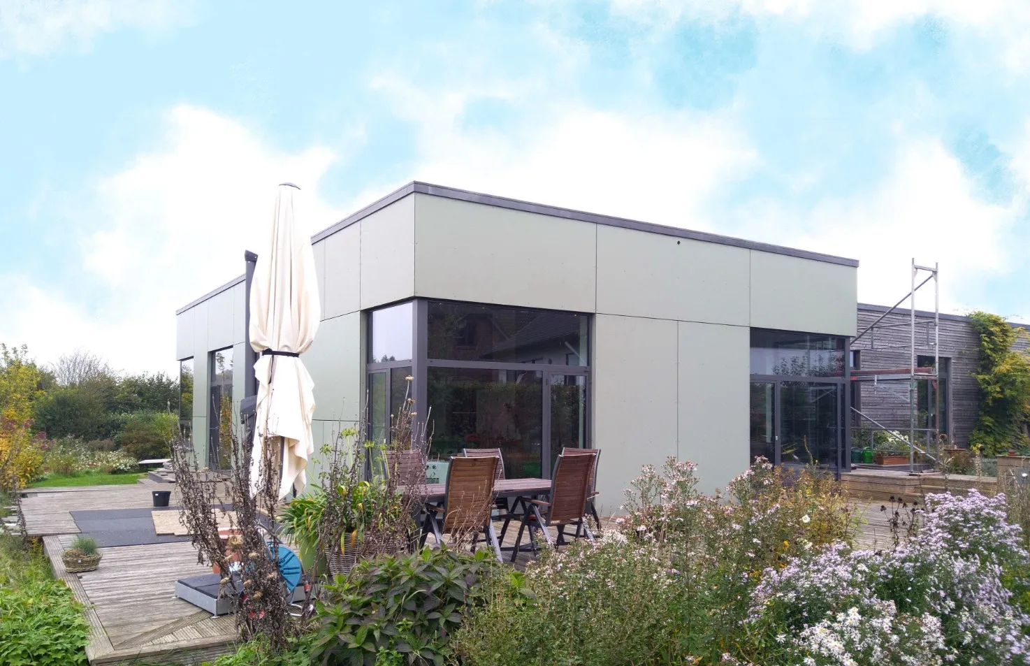 Modern flaches Haus mit großer Terrasse, Garten und Gartenmöbeln unter blauem Himmel.