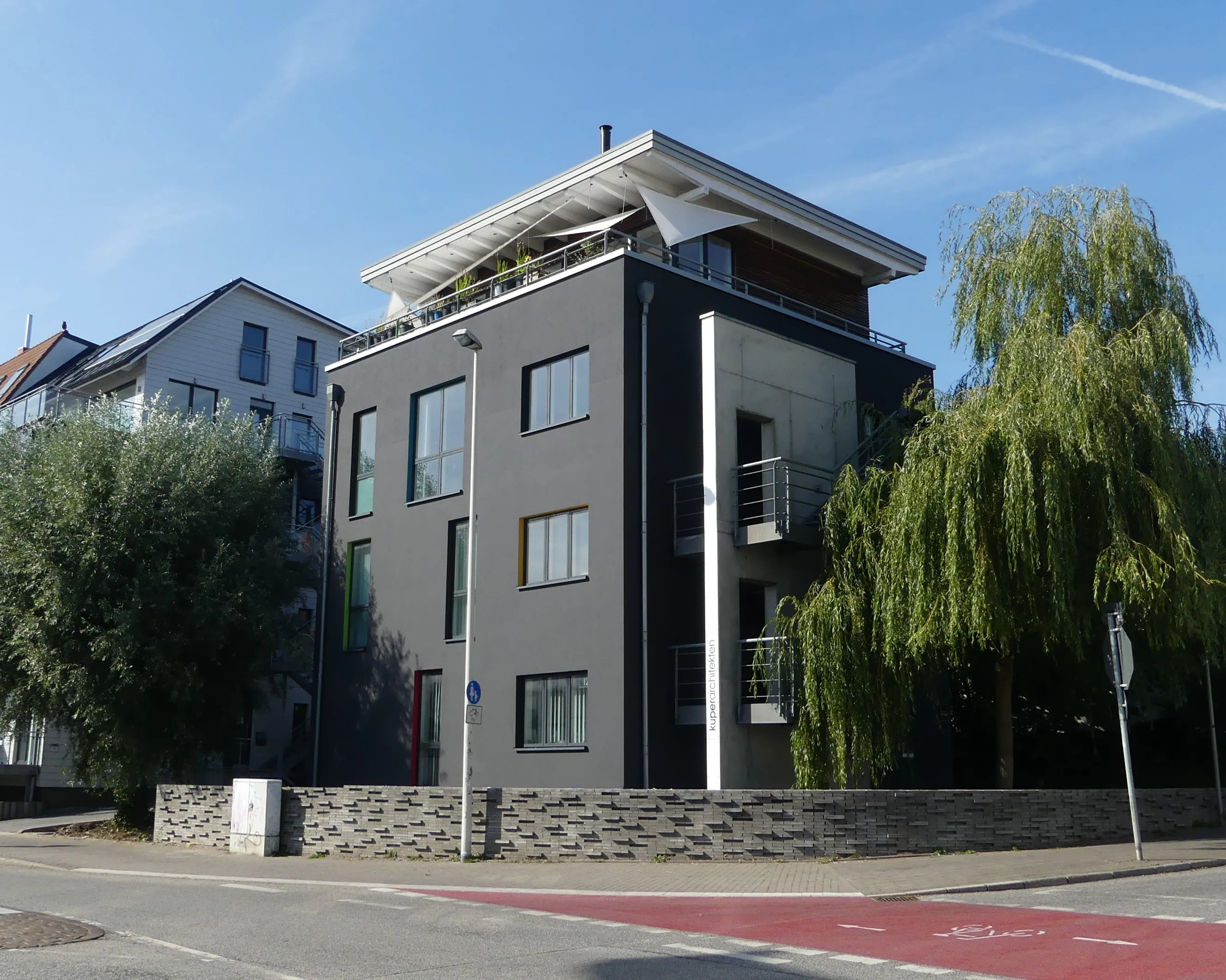 Modern dark gray building with large windows, balcony, and trees on a sunny day.