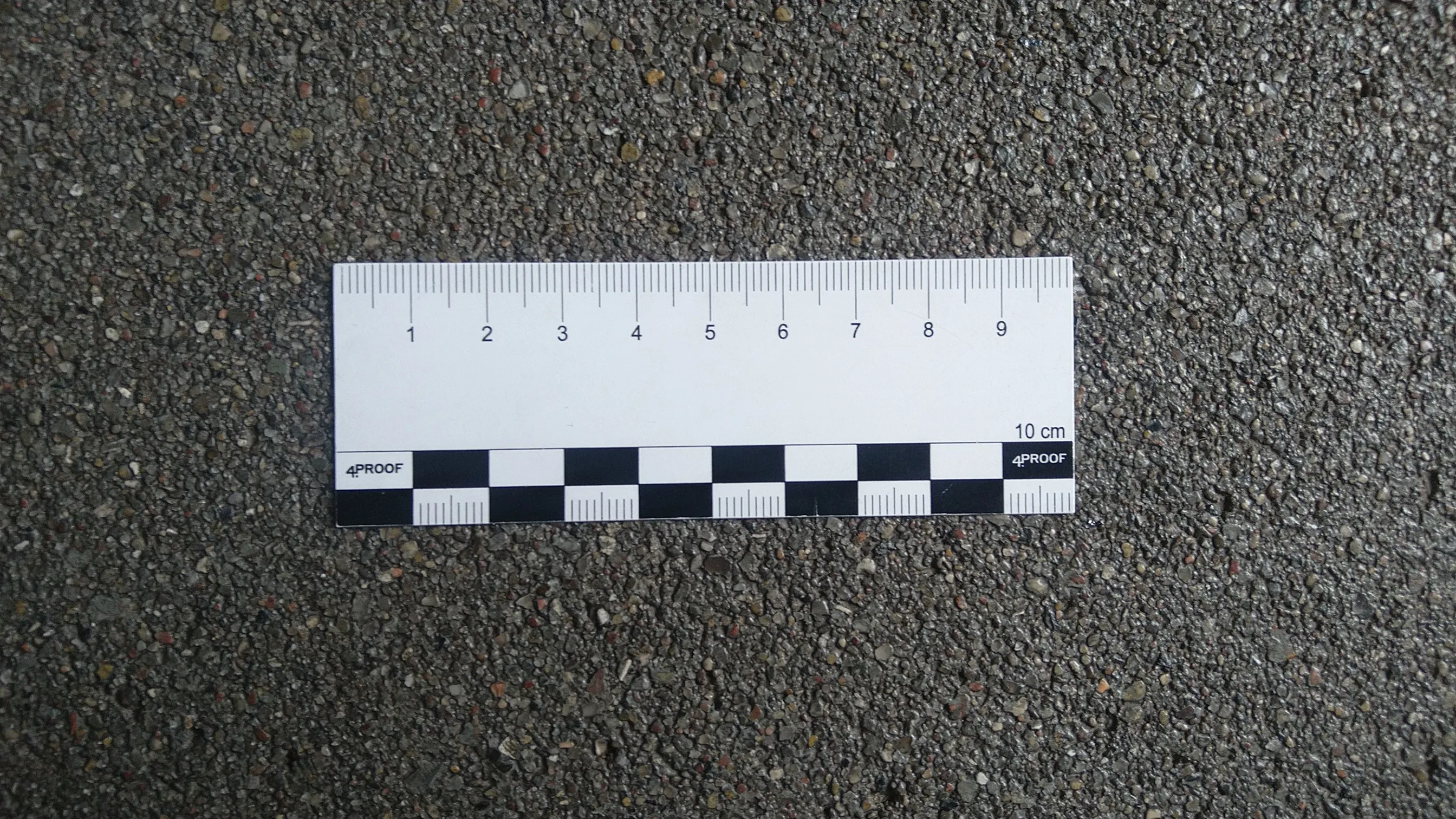 Weiße Messleiste mit Zentimetereinheiten und schwarz-weißen Kontrollfeldern auf grau körnigem Boden.