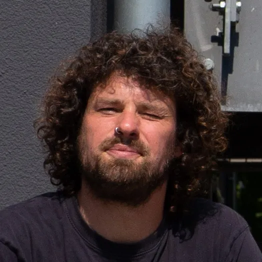 Mann mit lockigem Haar und Bart trägt schwarzen Pullover und blickt zur Kamera im Sonnenlicht.