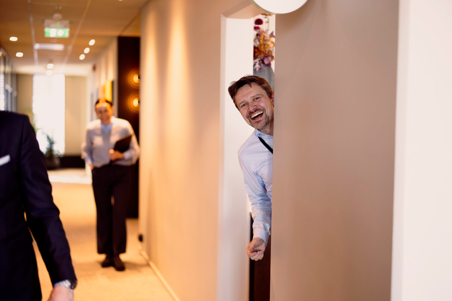 Smilende mann som kikker rundt hjørnet i en kontorgang med andre ansatte i bakgrunnen.