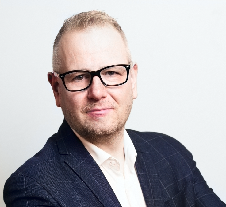 Mann med kort lyst hår, briller og mørk dressjakke smiler lett mot kameraet.