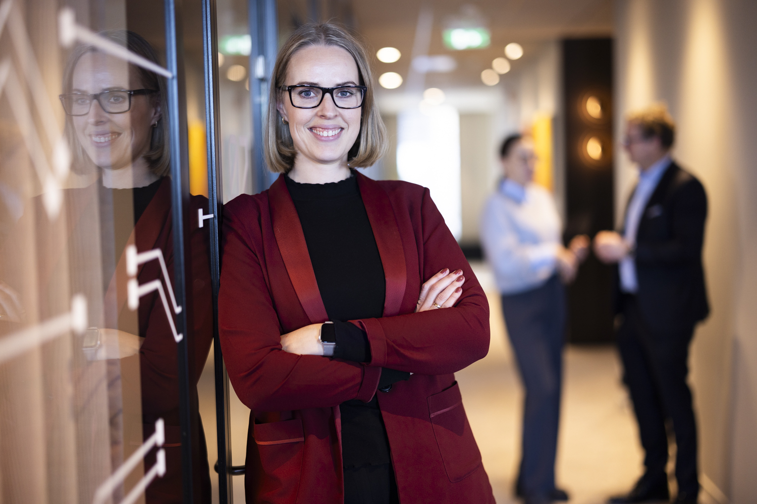 Smilende kvinne i rød blazer med armer krysset står ved en glassvegg i et kontor, med to personer i bakgrunnen som snakker sammen.