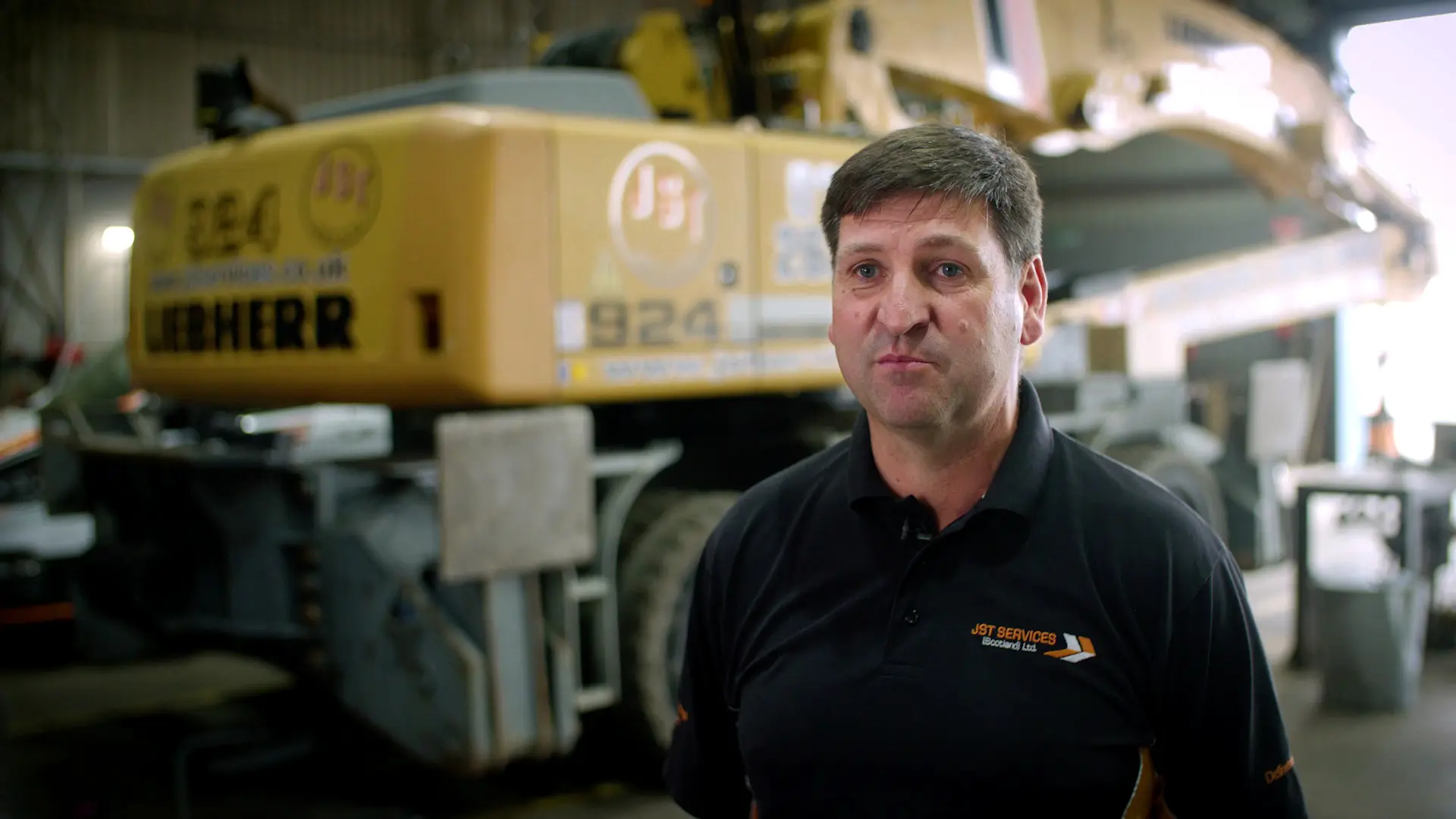 A man wearing a black shirt stands in front of a yellow crane.