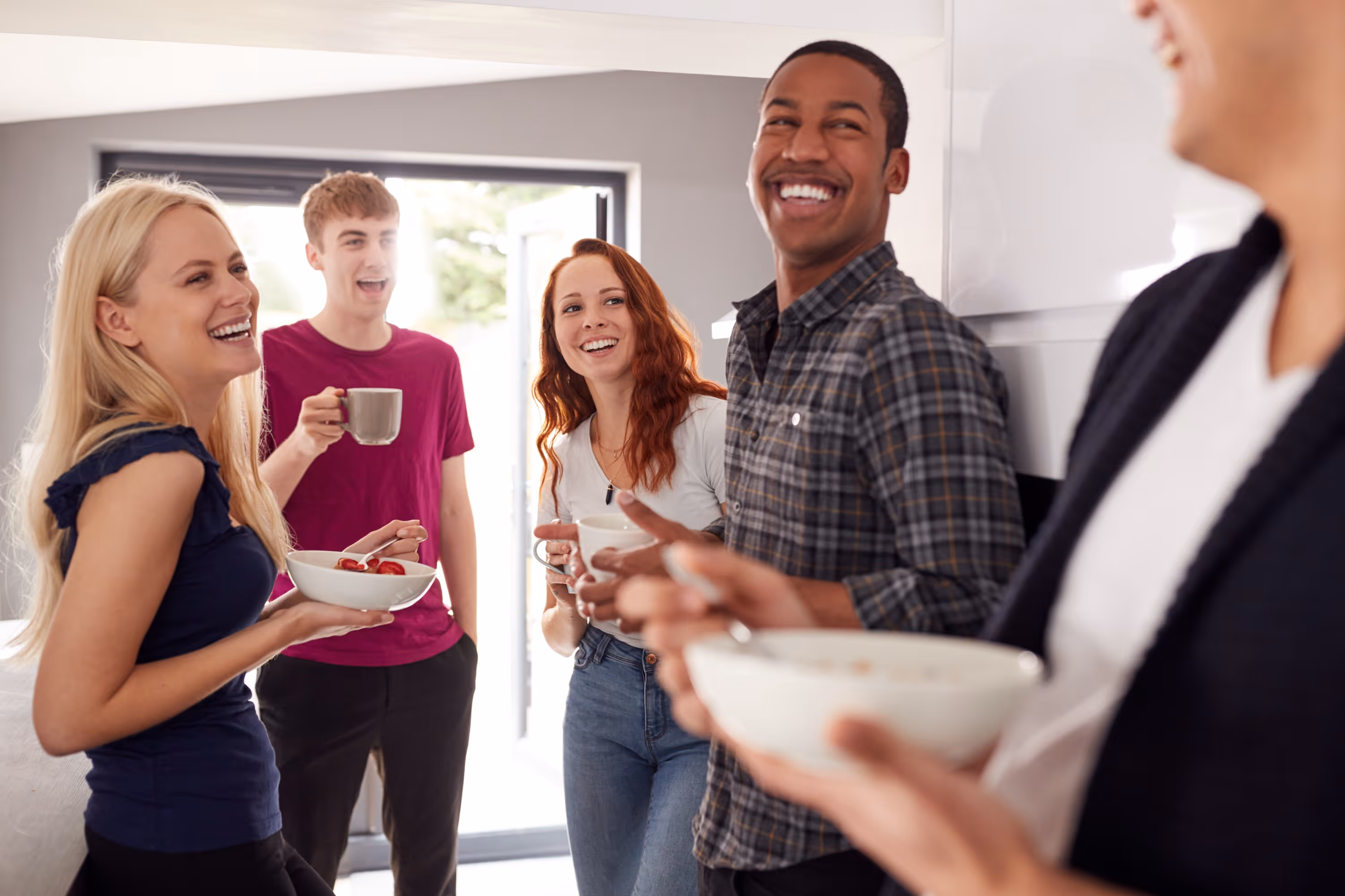 A group of four smiling roommates or friends laughing and socializing while holding coffee mugs and breakfast bowls in a bright, modern apartment kitchen at ArtHaus Dwight in Berkeley, CA.
