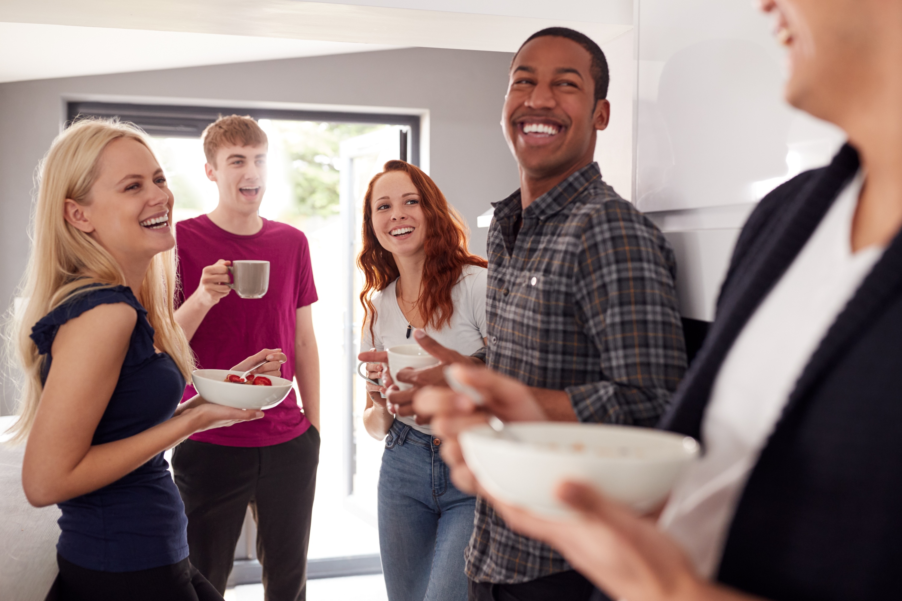 A group of four smiling roommates or friends laughing and socializing while holding coffee mugs and breakfast bowls in a bright, modern apartment kitchen at ArtHaus Dwight in Berkeley, CA.