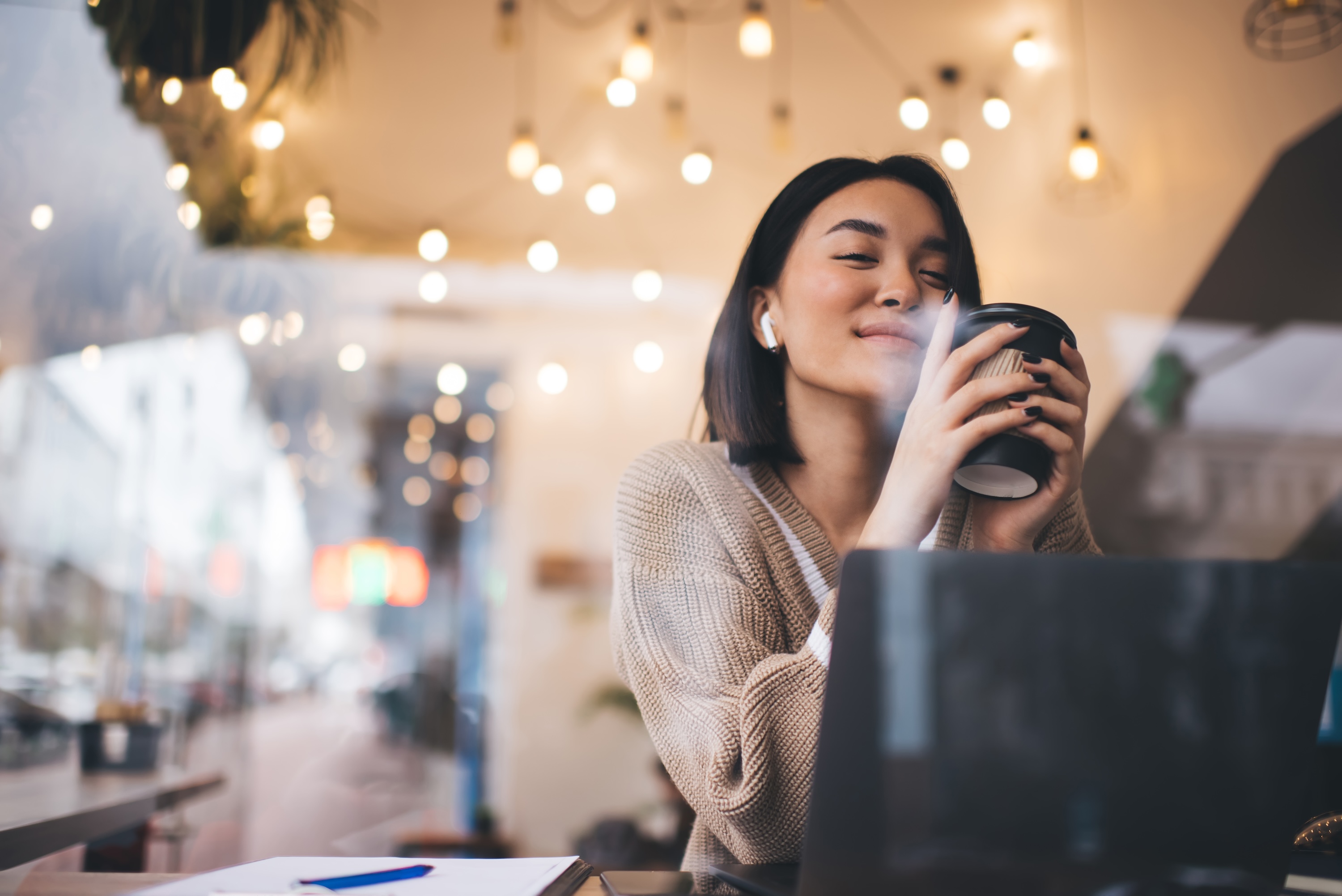 Woman in a cafe in Berkeley with a laptop, smiling with her eyes closed while holding a warm coffee cup.