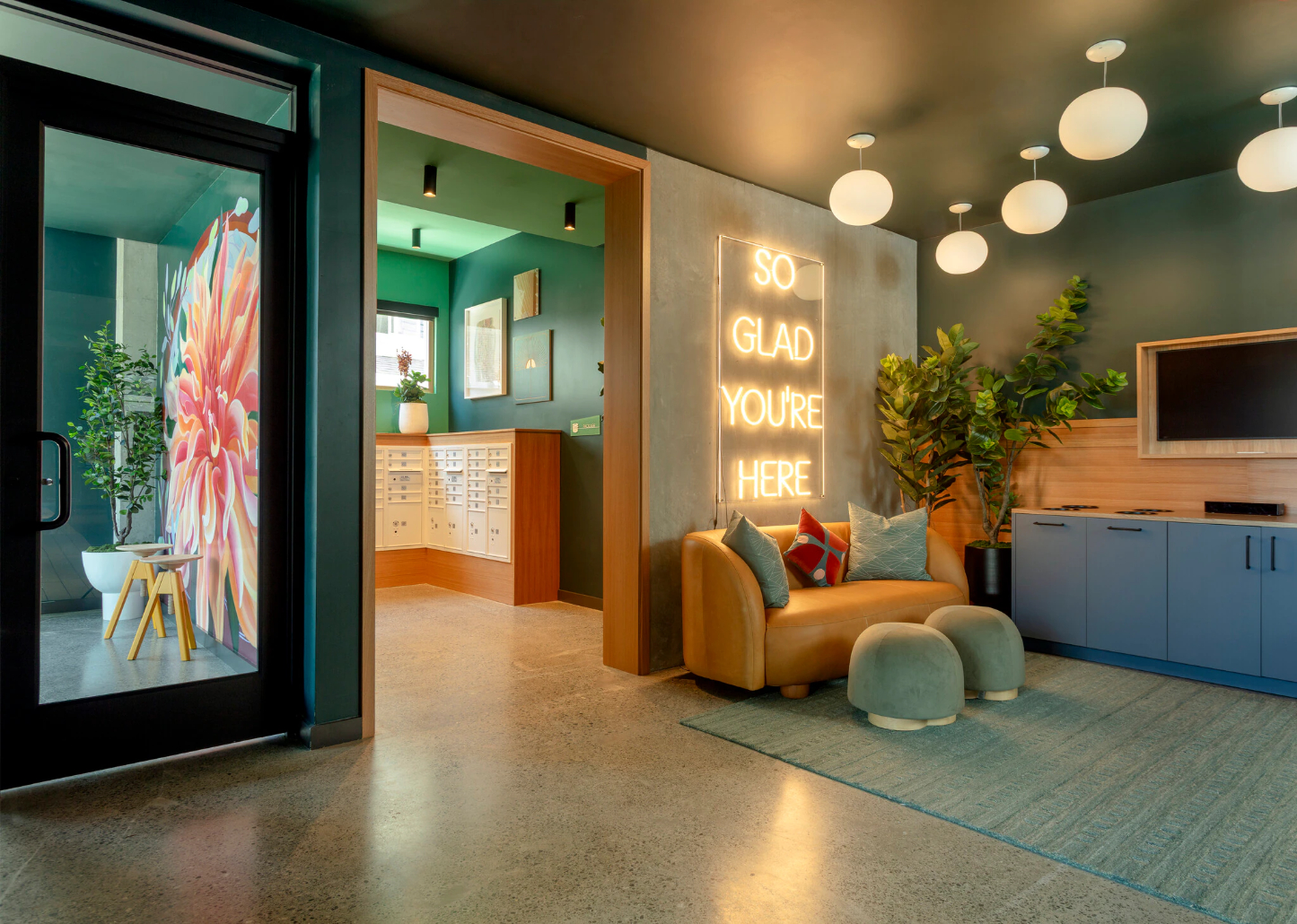 A modern resident common area with a television screen, a neon sign that says "SO GLAD YOU'RE HERE," and comfortable orange seating, set against accent walls with wood paneling and dark teal trim.