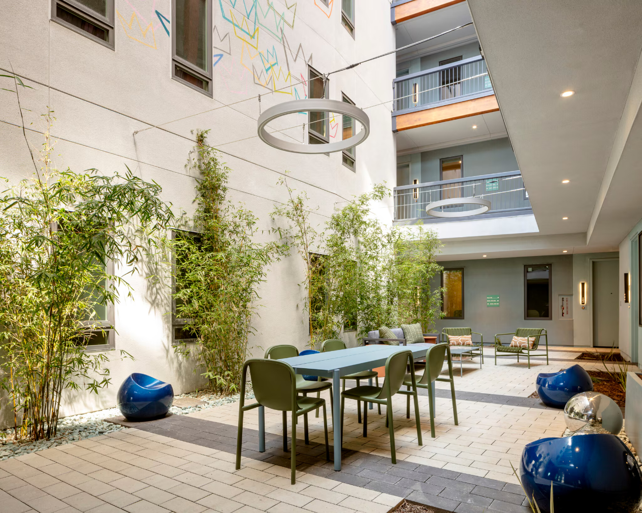 A landscaped outdoor courtyard area featuring a paved patio, a dining table with chairs, comfortable outdoor seating, and tall bamboo plants along the edge with string lights overhead.
