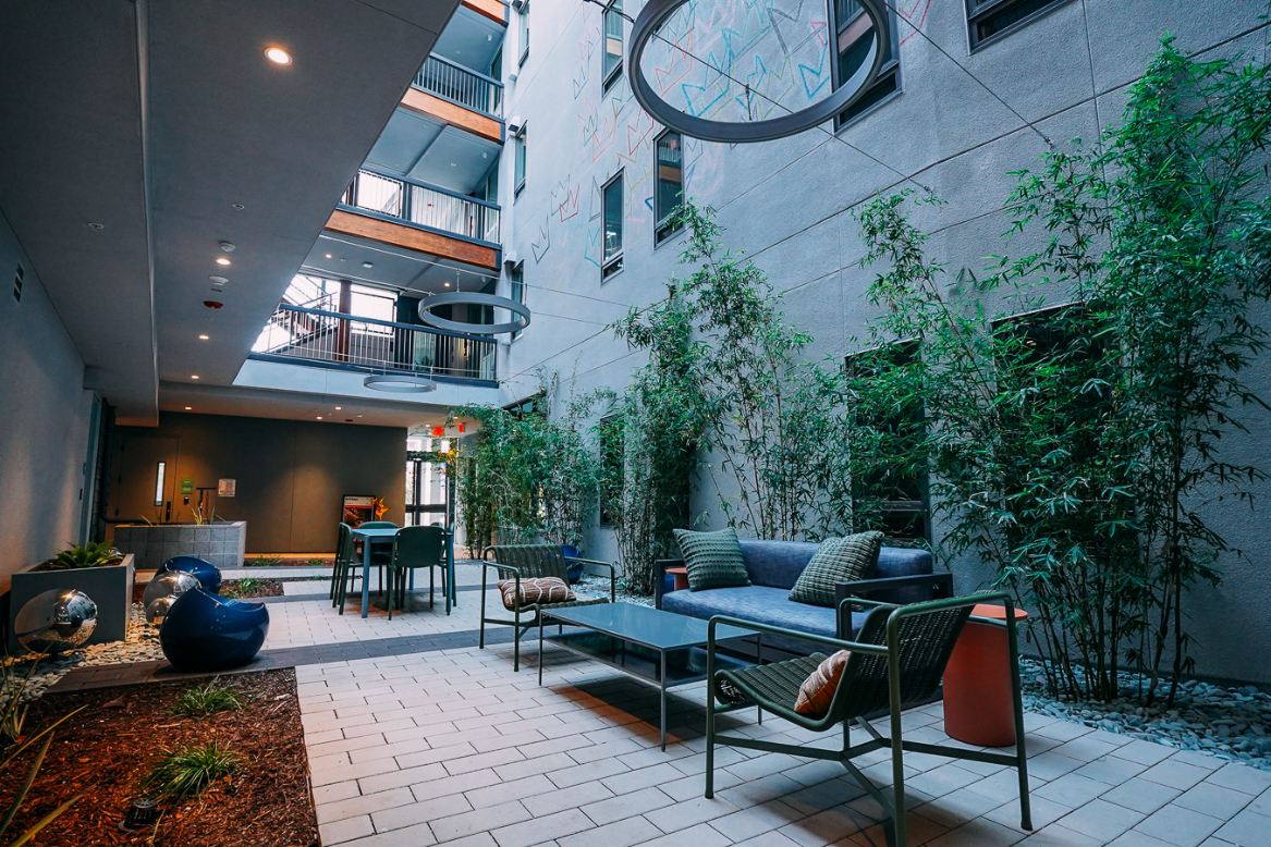 A ground-level view of the exterior courtyard with a modern outdoor sofa and side table in the foreground, facing a dining area nestled between bamboo dividers and landscaping.