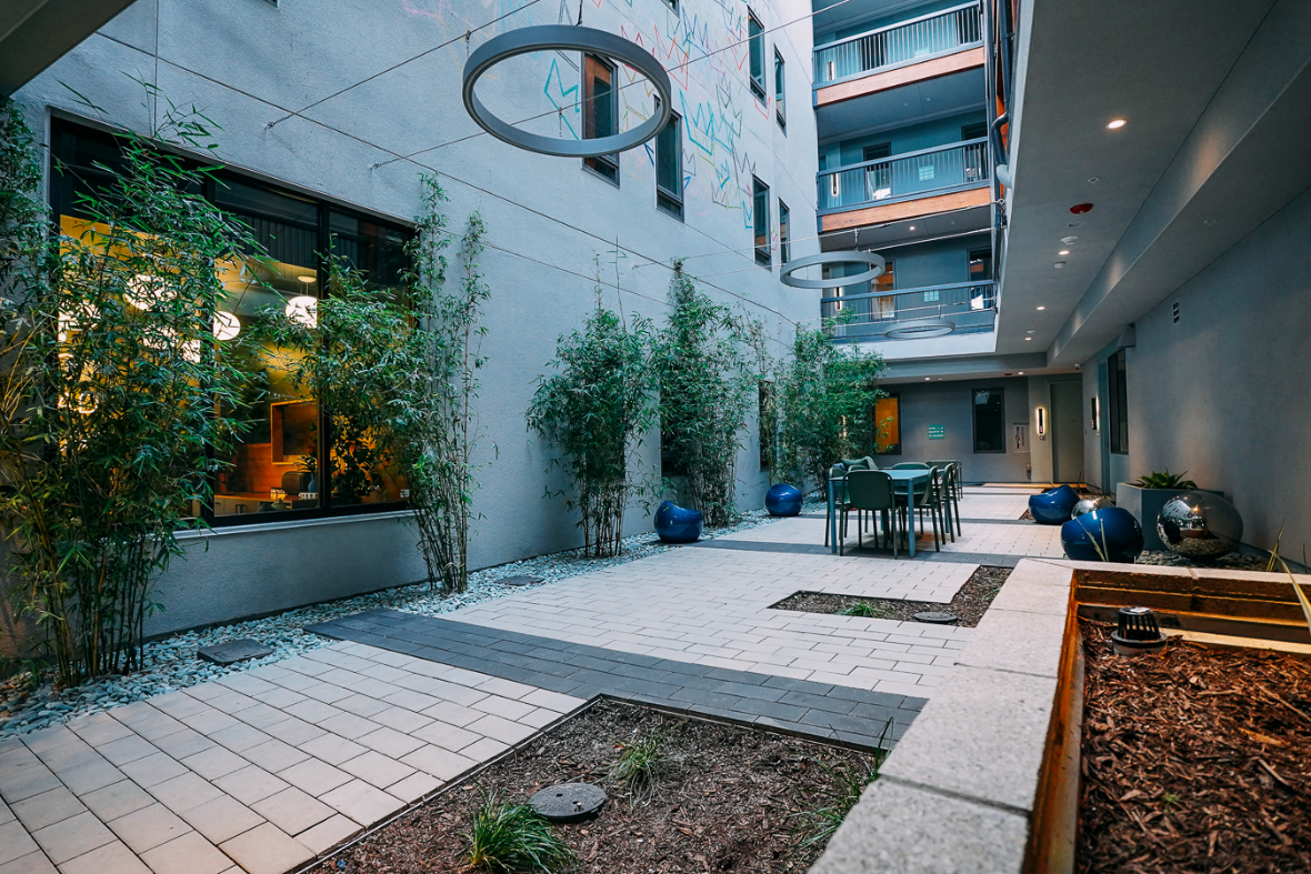 An outdoor, landscaped courtyard area featuring string lights overhead, a paved patio with dining furniture, and bamboo plants. The open-air apartment corridors are visible above.