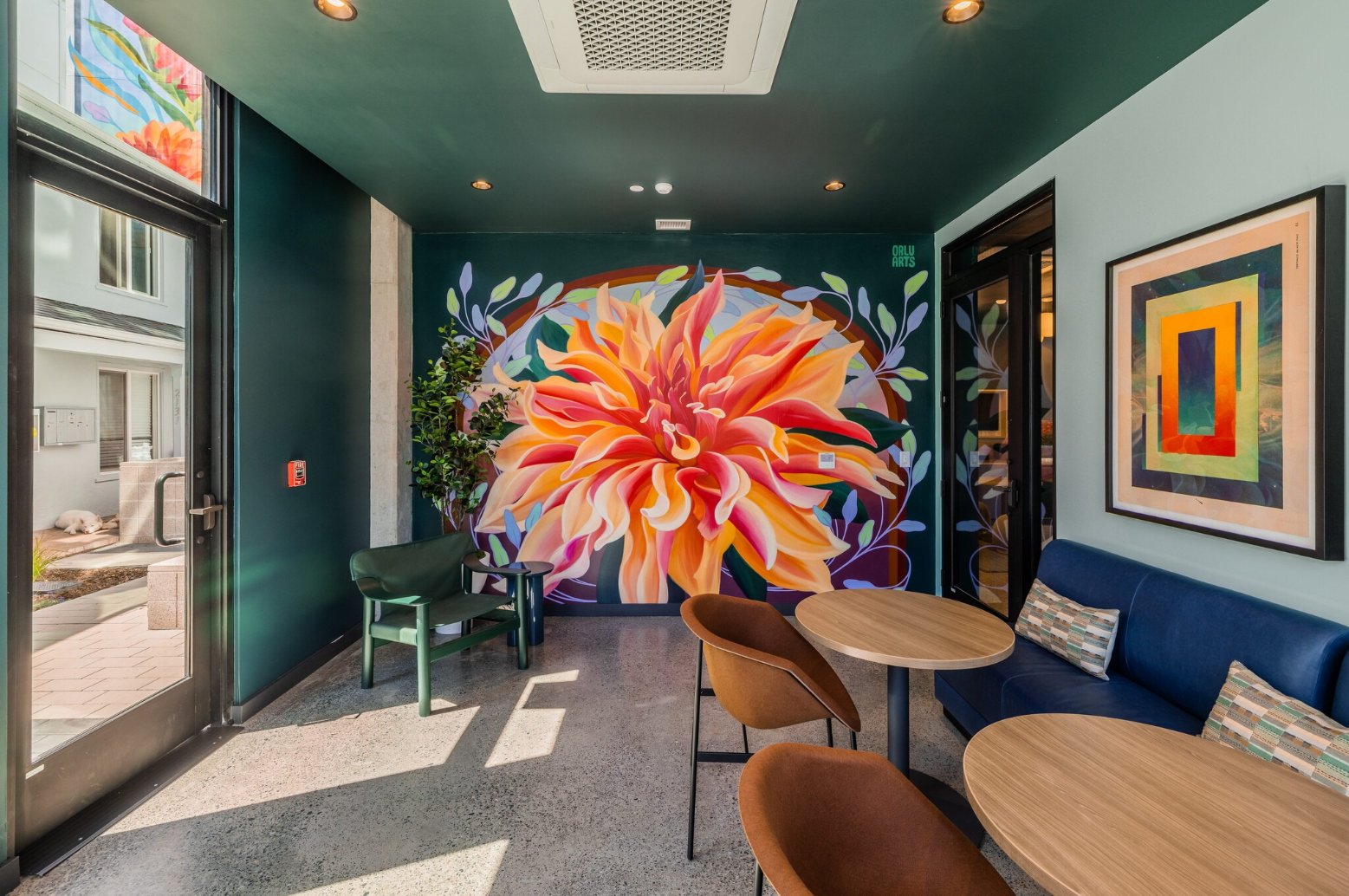 The indoor community lounge with floor-to-ceiling windows offering views of the exterior. The space features a long navy blue built-in couch, wooden tables, and a breakfast bar with modern stools.