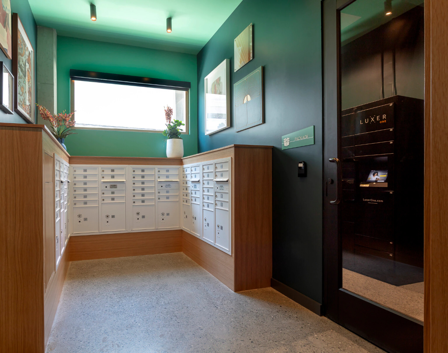 The community mailroom, featuring multi-tiered mail boxes, package locker, and a colorful, green back wall with flowers..