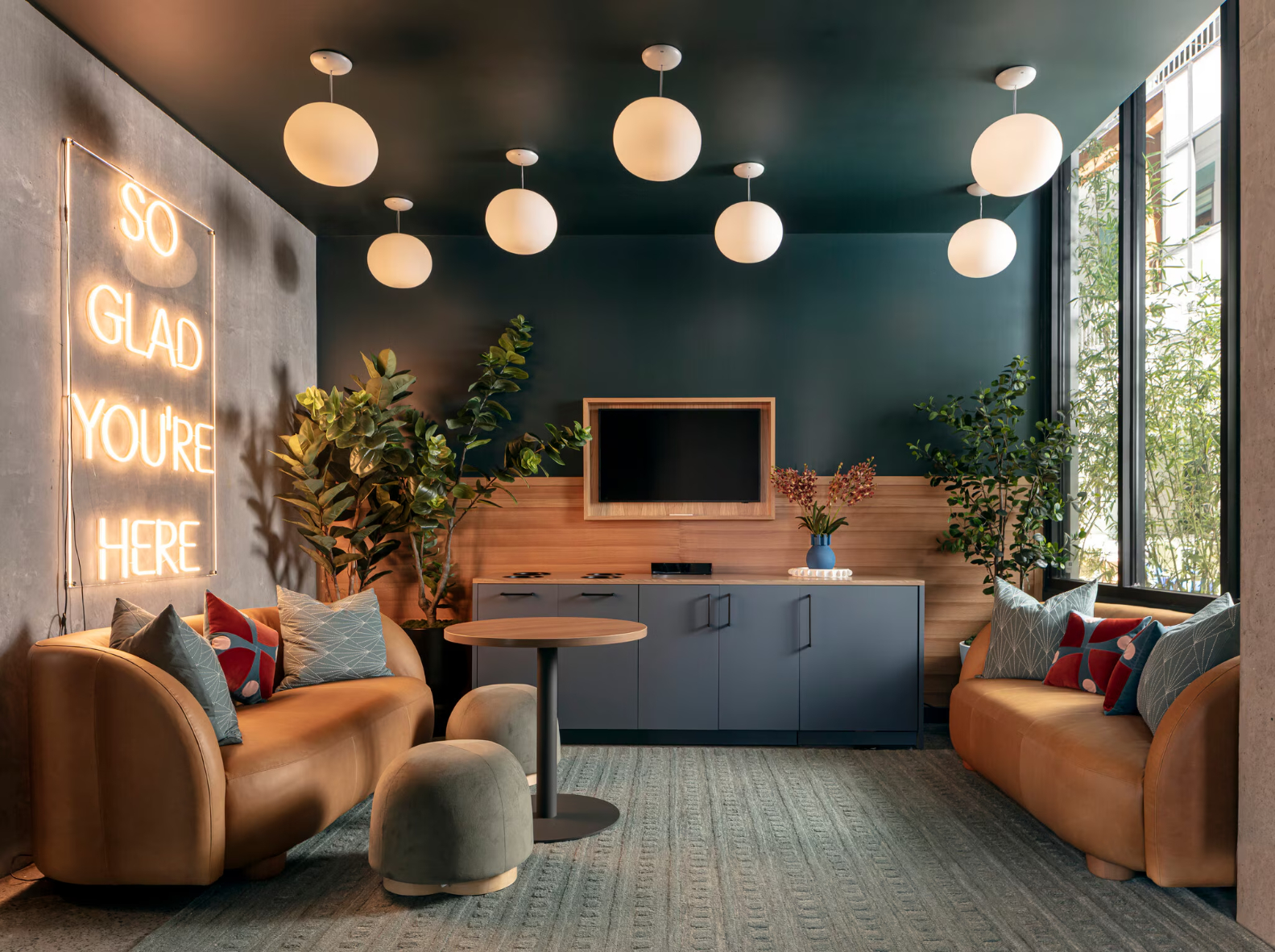 A modern resident common area with a television screen, a neon sign that says "SO GLAD YOU'RE HERE," and comfortable orange seating, set against accent walls with wood paneling and dark teal trim.