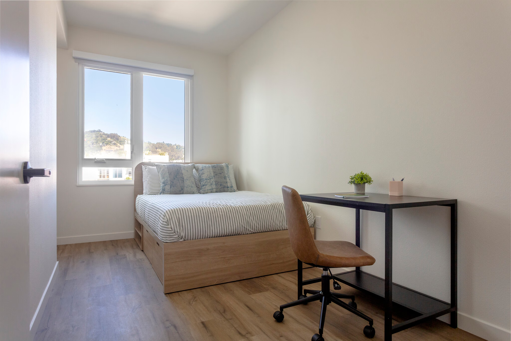 Fully furnished apartment bedroom featuring a bed with built-in storage drawers, a dedicated desk area, and large windows with blinds at ArtHaus Dwight in Berkeley, CA.