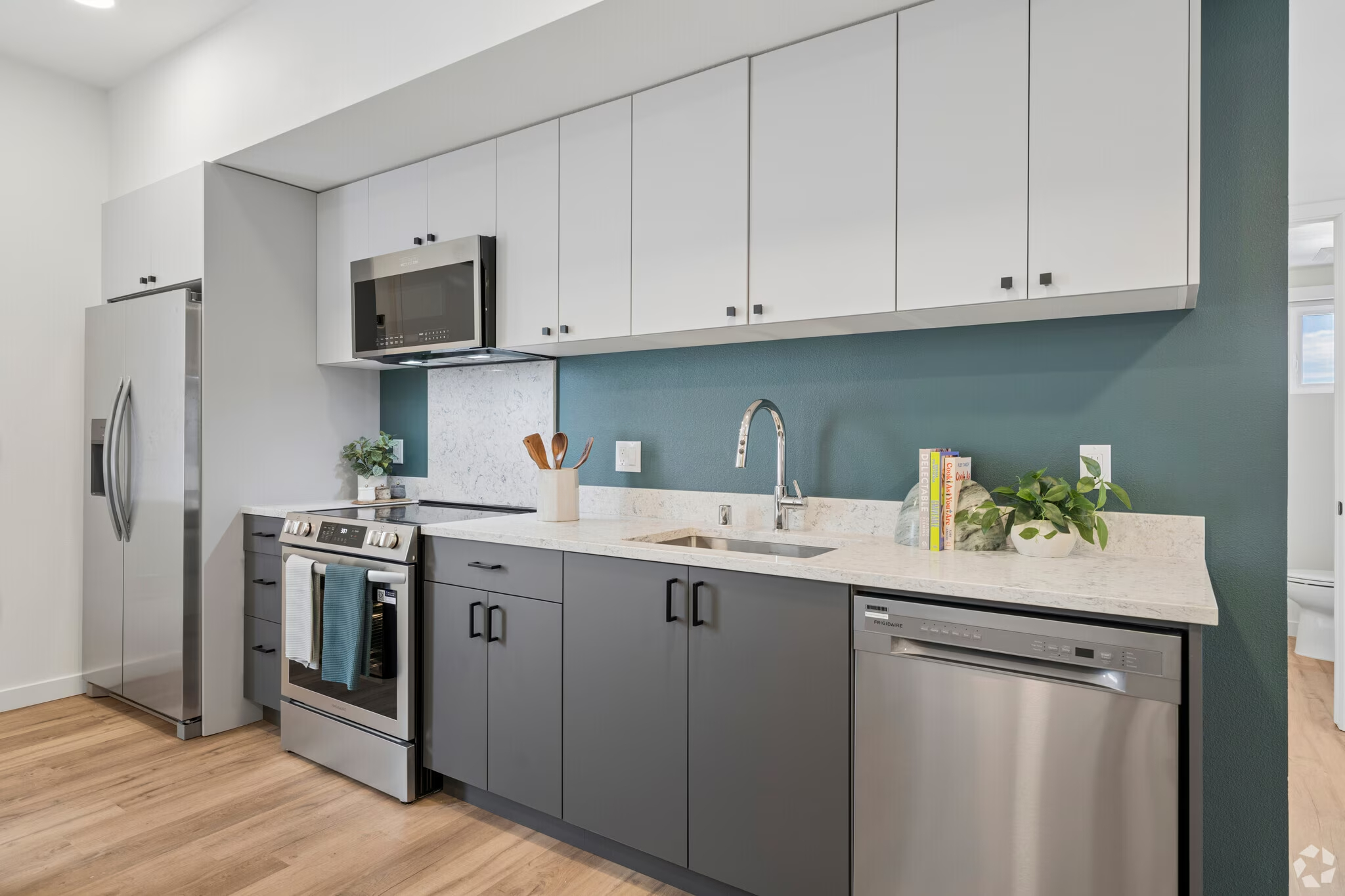 Modern apartment kitchen with two-tone cabinetry (green base, white upper), stainless steel appliances, and a leather sofa in the adjacent living area at ArtHaus Dwight in Berkeley, CA.