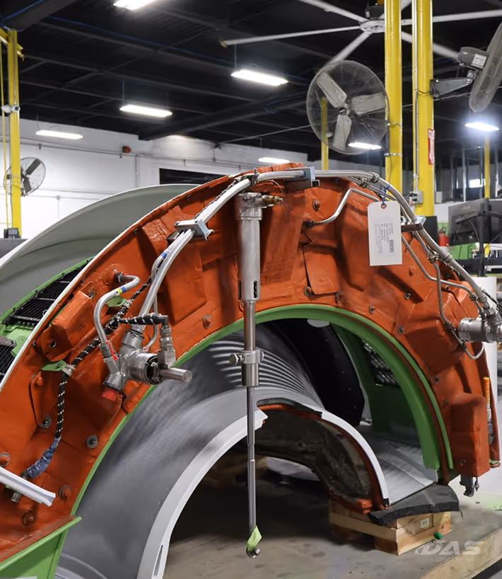 Mechanical assembly of a large industrial component with orange and green parts connected by metal pipes and mounted on a wooden pallet in a factory setting.