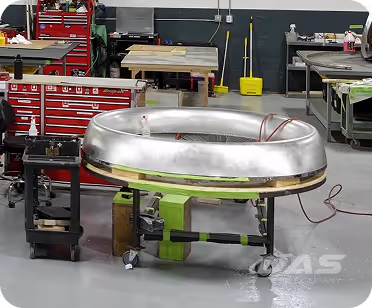 Large metallic toroidal object on a green stand with wheels inside an industrial workshop.