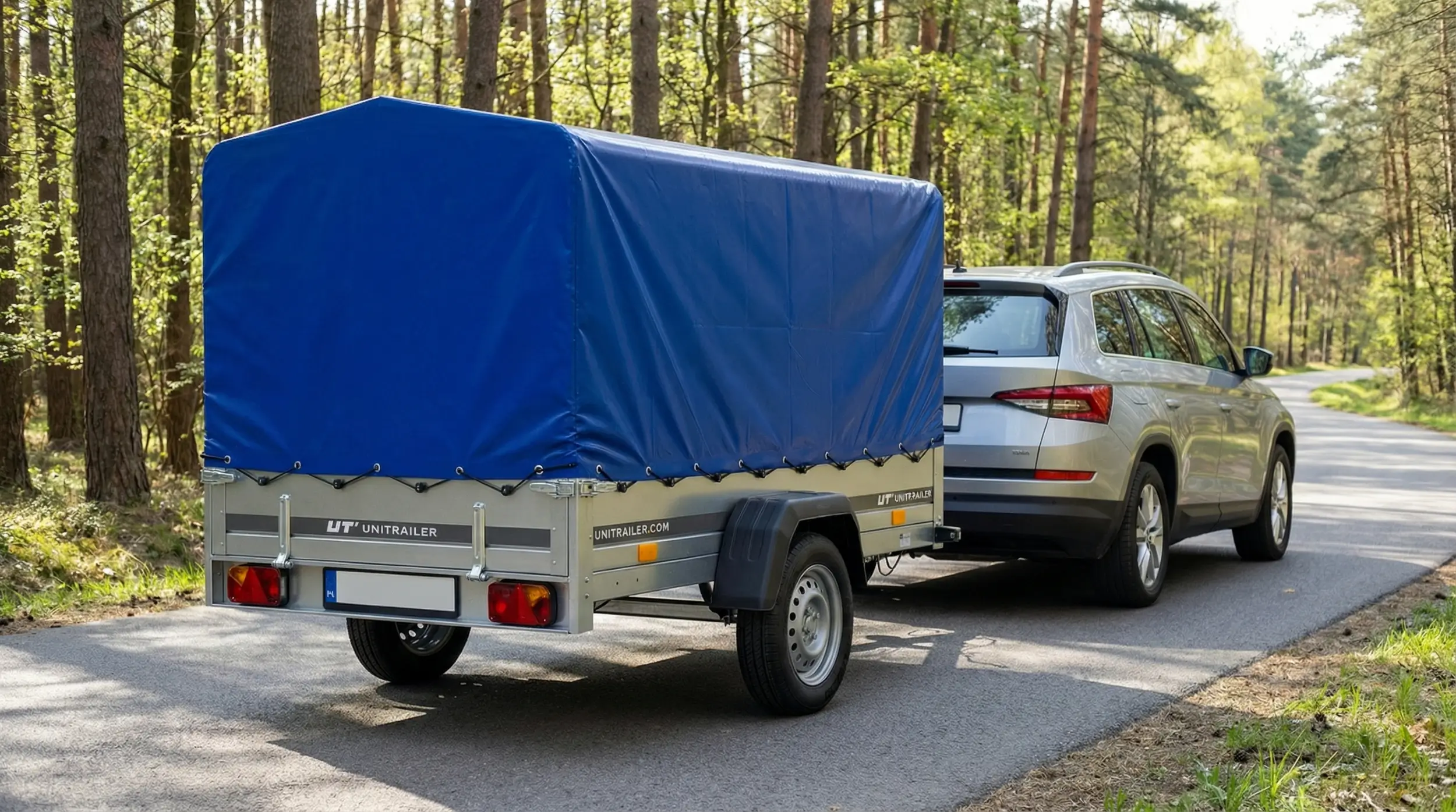 Przyczepka samochodowa Unitrailer Garden 230 FD z niebieską plandeką i załadowanymi rowerami, podpięta do srebrnego SUV-a na leśnej drodze.