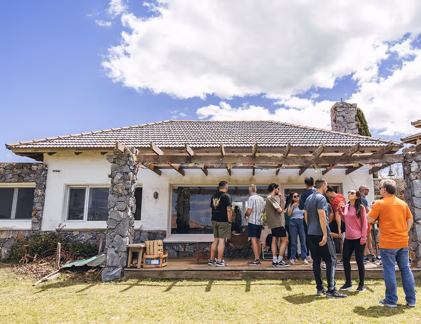 Group of people socializing on the porch of a stone and tile-roofed house under partly cloudy sky.